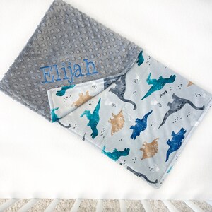 May include: A gray and white baby blanket with a dinosaur print on one side and a gray minky dot fabric on the other side. The blanket is personalized with the name "Elijah" embroidered on the gray side.