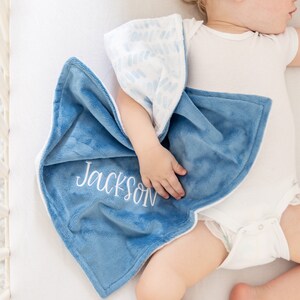May include: A soft, blue baby blanket with the name "Jackson" embroidered in white. The blanket has a plush texture and a white border. The reverse side features a white fabric with a blue pattern. The blanket is draped over a baby.