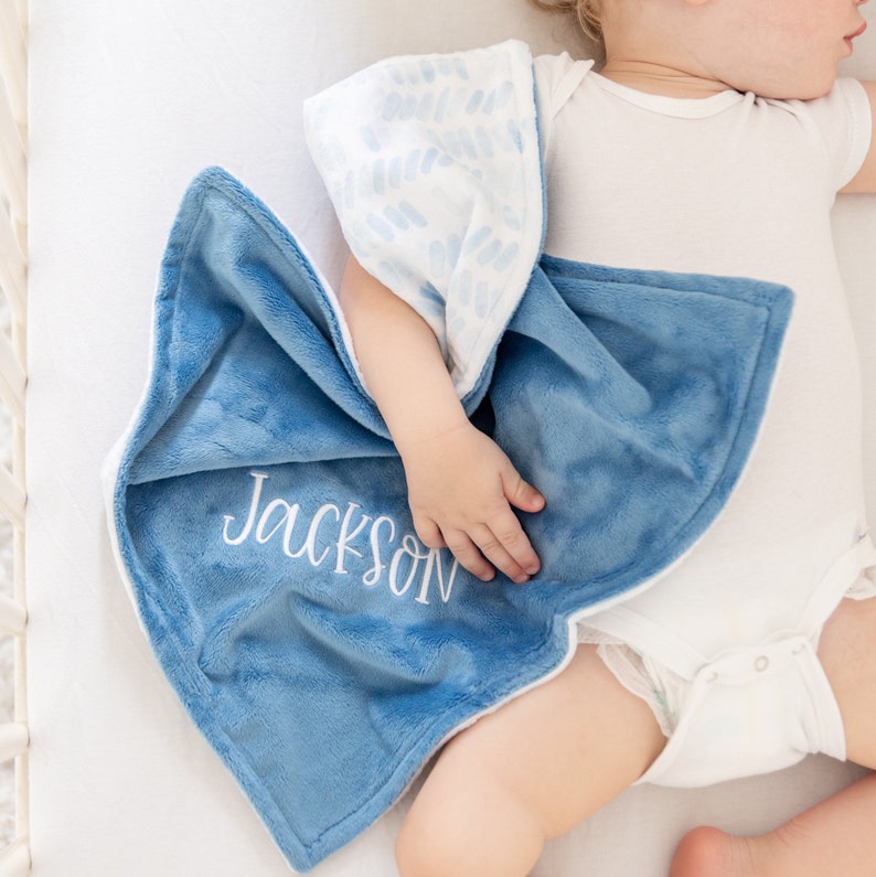 May include: A soft, blue baby blanket with the name "Jackson" embroidered in white. The blanket has a plush texture and a white border. The reverse side features a white fabric with a blue pattern. The blanket is draped over a baby.
