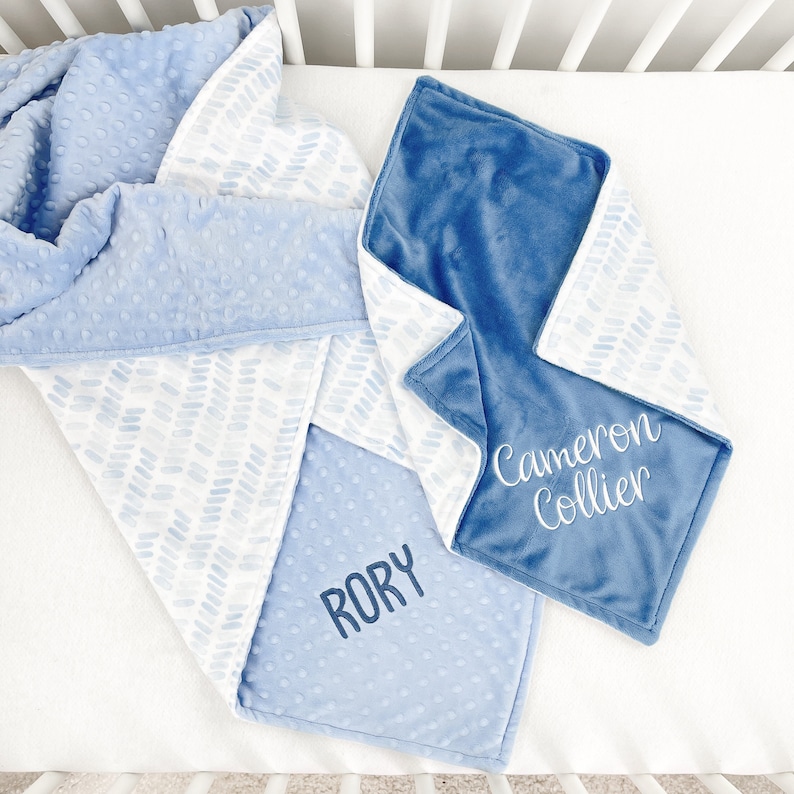May include: Two baby blankets in shades of blue. One blanket has a textured, dotted pattern, while the other features a chevron design. The name "RORY" is embroidered on one blanket, and "Cameron Collier" on the other.