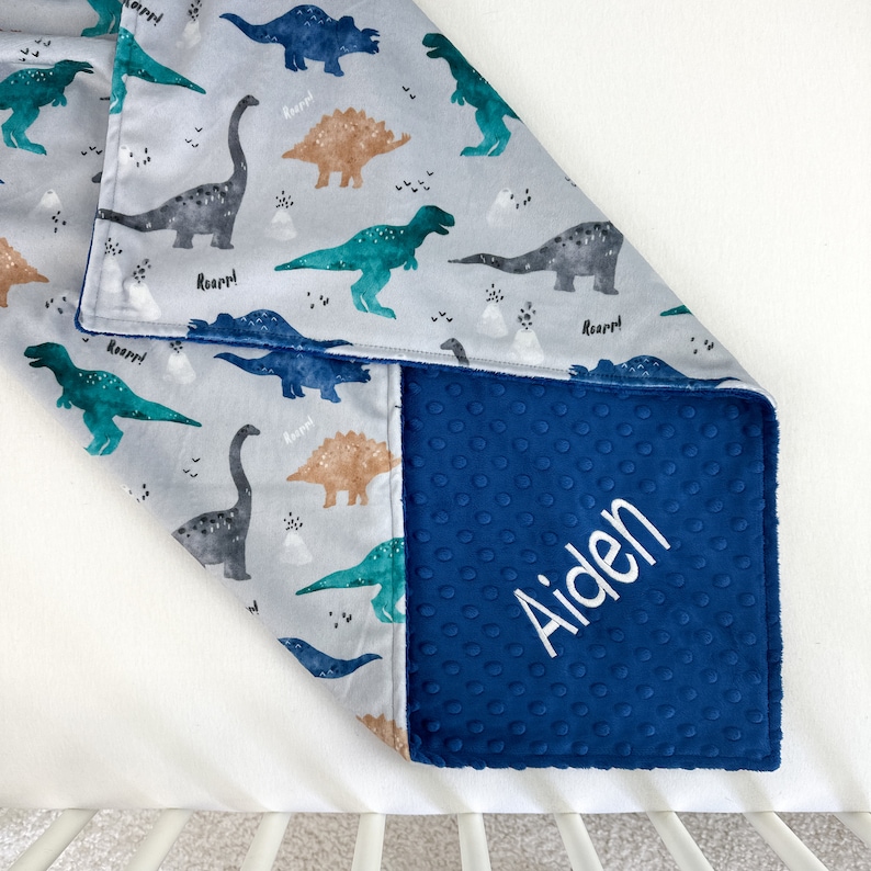May include: A blue and gray baby blanket with a dinosaur print and the name "Aiden" embroidered on the blue side.