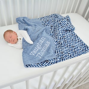 May include: A baby blue blanket with the name "Vaughan Thomas" embroidered in white. The blanket has a textured, dotted surface and is paired with a patterned blanket in shades of blue and white. The blankets are on a white crib.