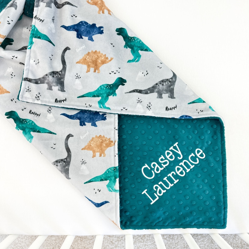 May include: A teal blue minky baby blanket with a gray and white dinosaur print on one side. The other side is solid teal blue with the name "Casey Laurence" embroidered in white.