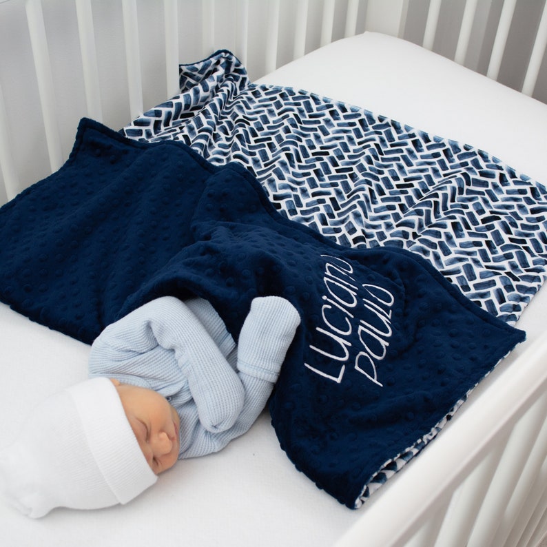 K&ouml;nnte beinhalten: Eine marineblaue Babydecke mit dem Namen "Luciano Paulo" in Wei&szlig; gestickt. Ein Baby ist in die Decke gewickelt und tr&auml;gt eine wei&szlig;e M&uuml;tze und einen hellblauen Strampler. Eine weitere Decke mit einem blau-wei&szlig;en geometrischen Muster ist ebenfalls zu sehen.