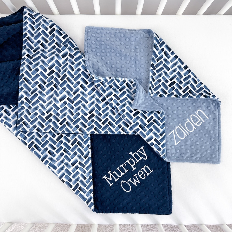 K&ouml;nnte beinhalten: Eine Patchwork-Babydecke mit einem marineblauen und wei&szlig;en Aquarell-Fischgr&auml;tenmuster, einem einfarbigen marineblauen Abschnitt und einem hellblauen Abschnitt. Die Decke ist mit den Namen "Zaidan" und "Murphy Owen" in Wei&szlig; bestickt.
