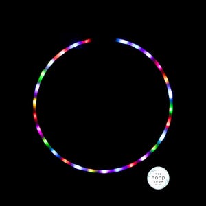 May include: A circular hula hoop illuminated with multicolored LED lights against a solid black background. The hoop features a continuous ring of glowing red, green, blue, and purple lights. A small circular logo reads The Hoop Shop est. 2014.