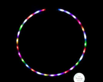 May include: A circular hula hoop illuminated with multicolored LED lights against a solid black background. The hoop features a continuous ring of glowing red, green, blue, and purple lights. A small circular logo reads The Hoop Shop est. 2014.