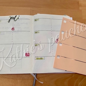 Op de afbeelding: Een weekplanner met het woord "Juli" bovenaan. De planner heeft een gestippeld rasterpatroon en is open op een pagina met een to-do-lijst. Een roze en witte sticker met een tasontwerp is op de pagina.