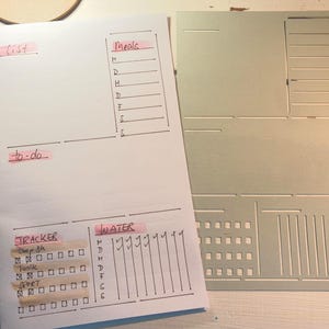 May include: A white paper planner with handwritten sections for lists, to-do items, and trackers. The planner includes sections for meals, water intake, and tracking sleep, work, and exercise. A gray template is next to the planner.