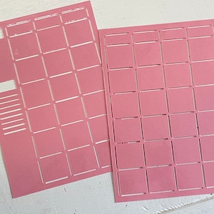 Könnte beinhalten: Zwei rosa Papierbögen mit einem Raster aus quadratischen Aussparungen. Ein Bogen hat zusätzliche rechteckige und linienförmige Aussparungen am linken Rand. Die Bögen liegen auf einer weißen Oberfläche. Die Aussparungen erzeugen ein Muster aus kleinen Quadraten.