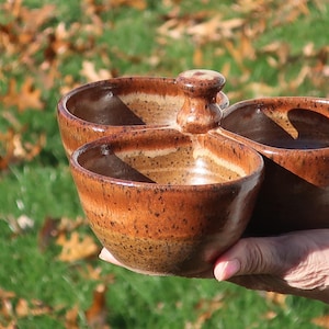 May include: A handmade, three-section ceramic serving dish with a rich brown glaze. The dish has a central handle and three separate bowls, perfect for dips, snacks, or condiments. The pottery is held in a hand against a blurred background of green grass and fallen leaves.