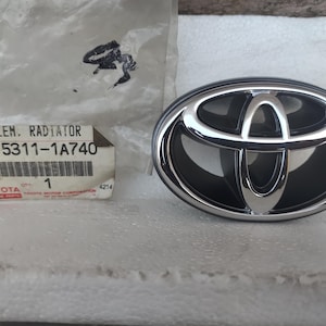 May include: A chrome and black Toyota radiator emblem. The oval-shaped emblem has a silver border and the Toyota logo. A small white label with text is also visible.