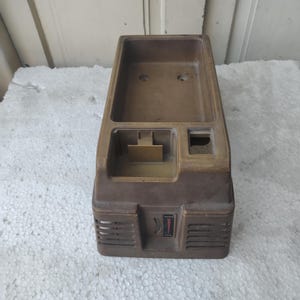 May include: A brown plastic housing for an electronic device featuring a large rectangular recessed compartment, a smaller square slot, and a front panel with horizontal ventilation slats and a small vertical indicator label with a red triangle.