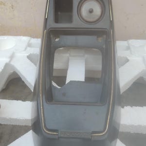 May include: A dark gray automotive center console trim piece featuring a rectangular storage compartment, a circular cutout for a gauge or clock, and a large central opening for a gear shifter. The word TOYOTA is embossed at the base.