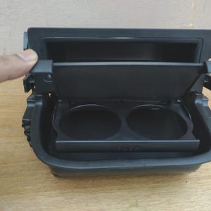 May include: A black plastic automotive cup holder assembly featuring a rectangular frame with a hinged lid that opens to reveal two circular recessed cup slots. The front edge of the cup holder tray has the word PUSH embossed in capital letters.