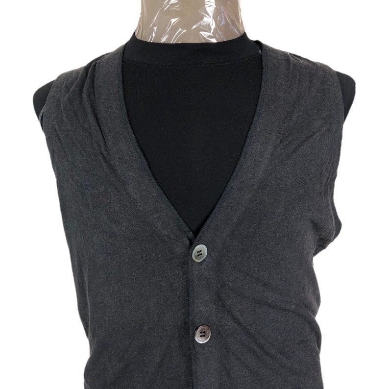 May include: A dark gray, button-down vest with a V-neck. The vest has two buttons and is displayed on a black mannequin. The vest is made of a soft, possibly knit, material.