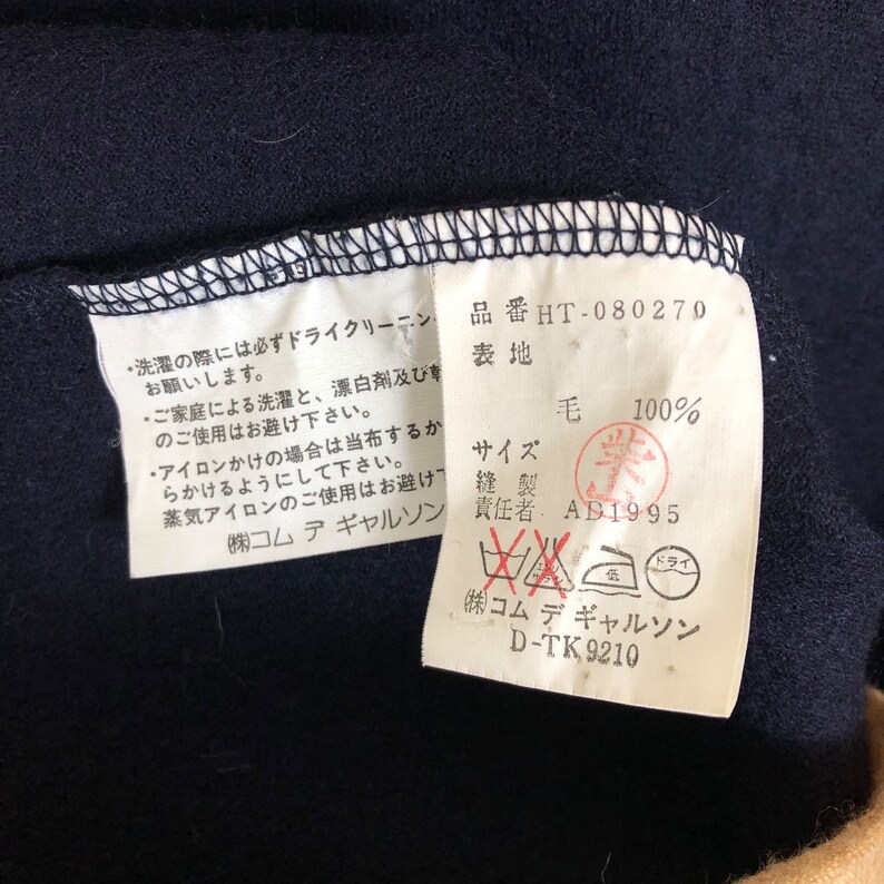 May include: Close-up of a navy blue fabric with attached tags. The tags contain Japanese text, care instructions, and a stamp. The fabric is 100% wool. The tag also includes the brand name and product code.