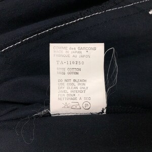 May include: Close-up of a black garment with a white label. The label reads "COMME des GARCONS MADE IN JAPAN" and includes care instructions. The fabric is 100% cotton.