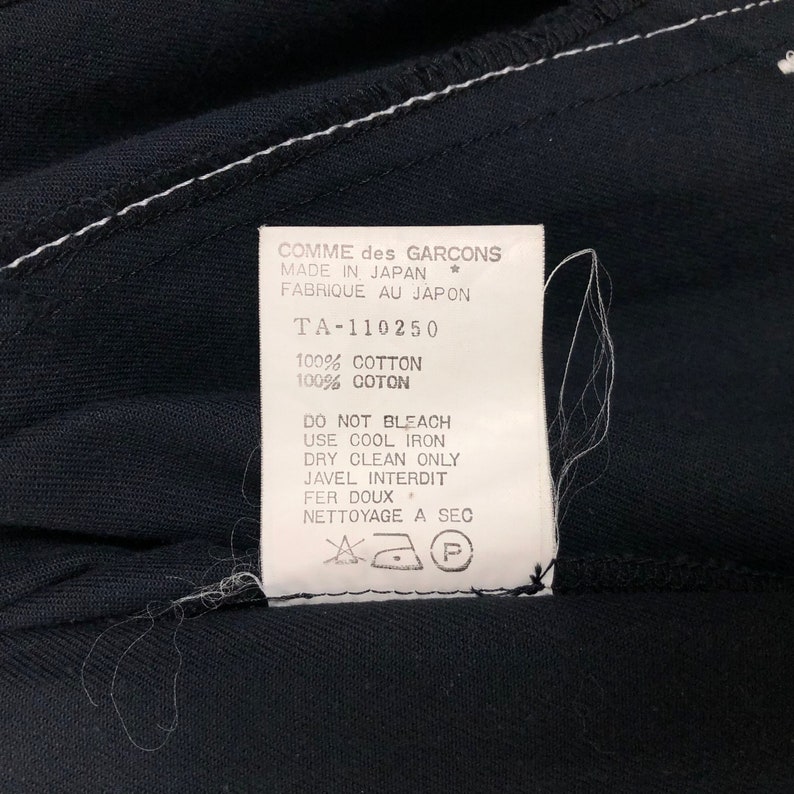 May include: Close-up of a black garment with a white label. The label reads "COMME des GARCONS MADE IN JAPAN" and includes care instructions. The fabric is 100% cotton.