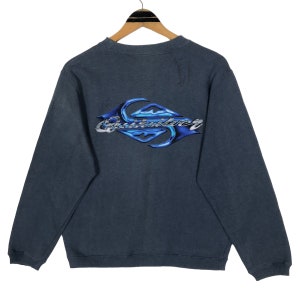 May include: A dark gray sweatshirt with a crew neck and long sleeves. The back features a blue and silver graphic of the word "Quiksilver" in a stylized flame design. The sweatshirt is hanging on a wooden hanger.