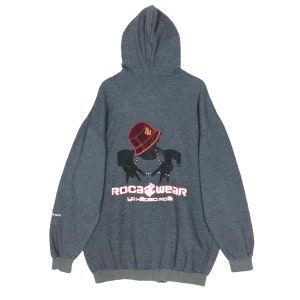 May include: A dark gray zip-up hoodie featuring a graphic on the back. The graphic includes a silhouette wearing a bucket hat and a necklace, with the text "ROCAWEAR" and "YO! HEEBO MOE" in red and white.