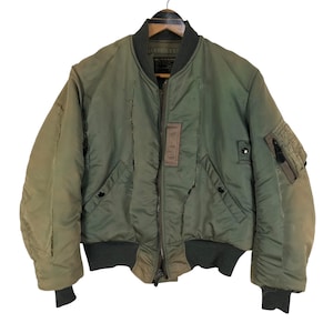 May include: Olive green bomber jacket with a ribbed collar, cuffs, and hem. The jacket features a zip-up front, two front pockets, and a patch on the chest. The jacket is hanging on a wooden hanger.