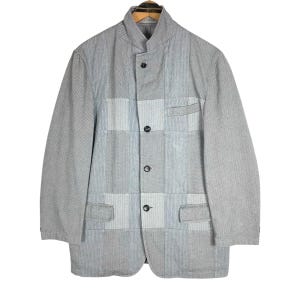 May include: A light grey jacket with a patchwork design. The jacket has a mandarin collar, three buttons, and two pockets. The fabric has a subtle striped pattern with blue accents. The jacket is hung on a wooden hanger.