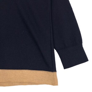 May include: Navy blue sweatshirt with a tan-colored bottom hem and sleeve cuffs. The long-sleeved top is made of a soft material. The image shows a close-up of the garment.