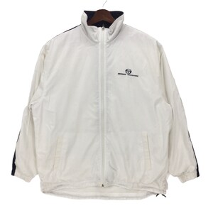 Puede incluir: Chaqueta blanca con cremallera, cuello y rayas laterales azul marino. La chaqueta presenta el logo "Sergio Tacchini" en el pecho. La chaqueta está colgada de una percha de madera.