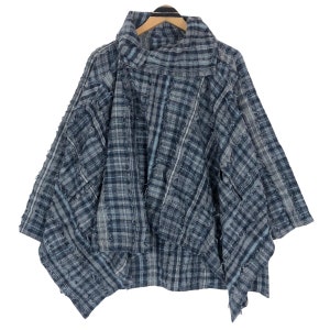Può includere: Un top a poncho a quadri blu e grigi con collo alto. Il tessuto presenta un design con bordi sfilacciati e usurati. L'indumento è appeso a una gruccia di legno su sfondo bianco.