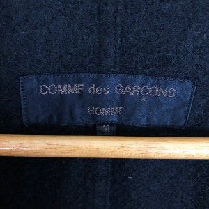 May include: Close-up of a black garment tag with the text "COMME des GARCONS HOMME" in a light color. The tag is rectangular and has a small star symbol. The size "M" is printed at the bottom. The tag is hanging on a wooden hanger.