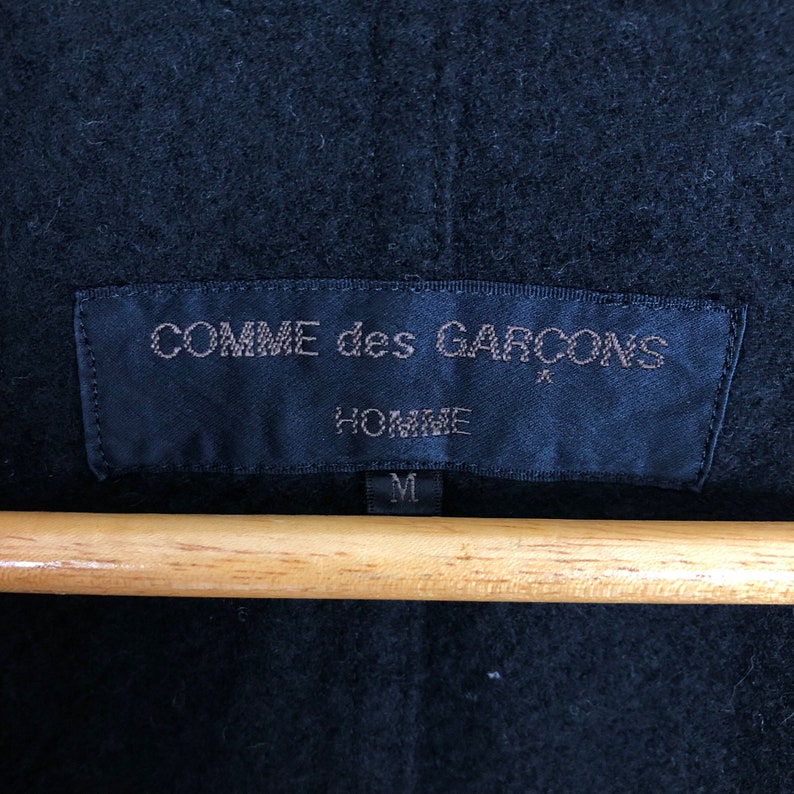 May include: Close-up of a black garment tag with the text "COMME des GARCONS HOMME" in a light color. The tag is rectangular and has a small star symbol. The size "M" is printed at the bottom. The tag is hanging on a wooden hanger.