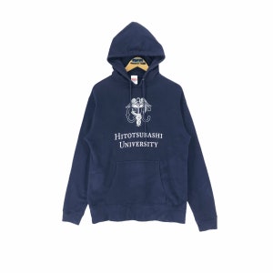 May include: Navy blue hooded sweatshirt with a front pocket and drawstring hood. The sweatshirt features a white graphic and the text "HITOTSUBASHI UNIVERSITY". The garment is displayed on a wooden hanger against a white background.
