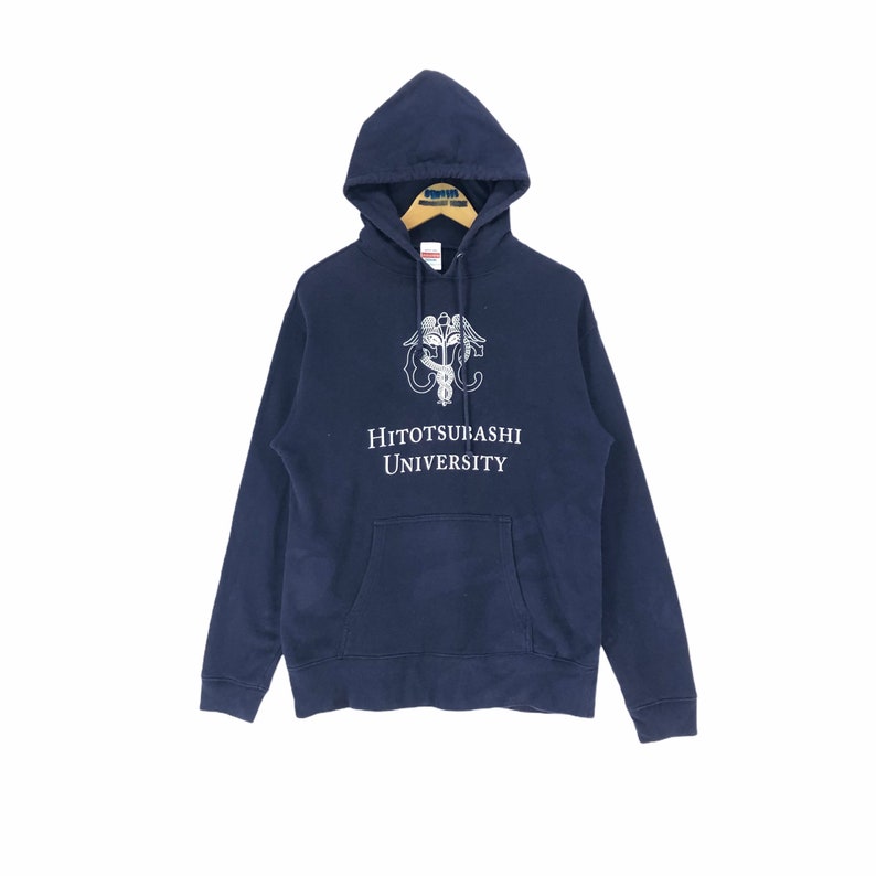 May include: Navy blue hooded sweatshirt with a front pocket and drawstring hood. The sweatshirt features a white graphic and the text "HITOTSUBASHI UNIVERSITY". The garment is displayed on a wooden hanger against a white background.