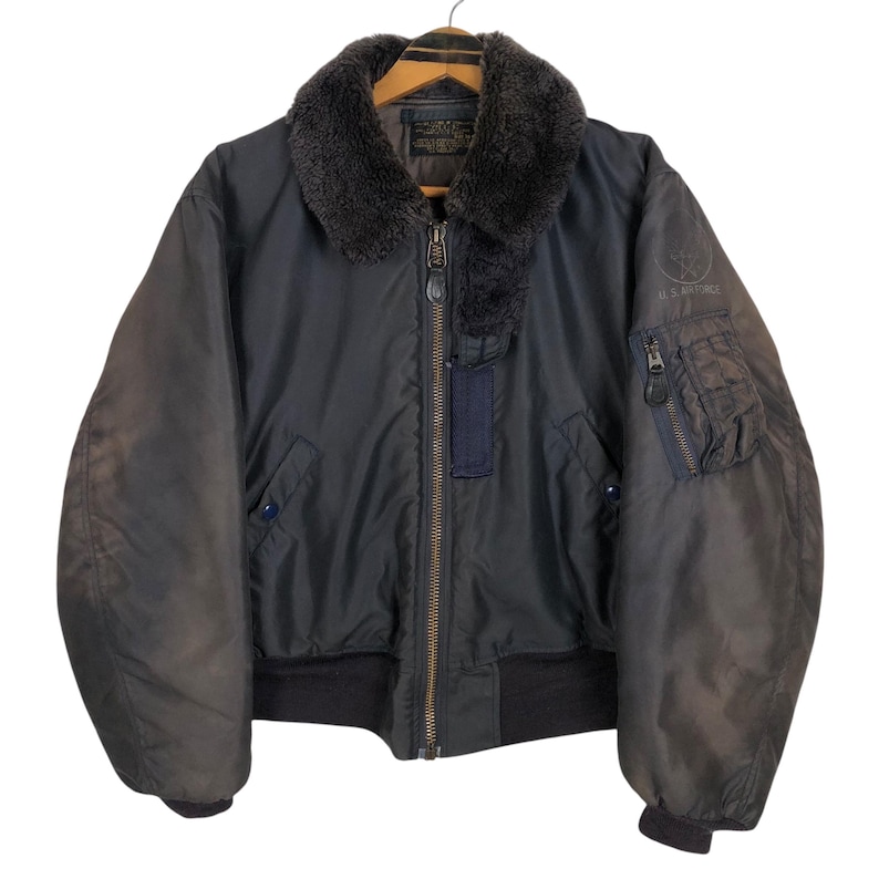 May include: A dark blue bomber jacket with a faux fur collar and a zipper closure. The jacket has a ribbed hem and cuffs, and a small pocket on the left sleeve. The jacket is hanging on a wooden hanger.
