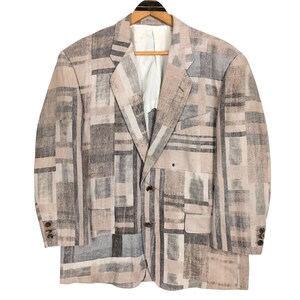 May include: A beige, gray, and black patterned blazer with a geometric design. The blazer has a notched lapel, two front pockets, and three buttons on each cuff. It is hanging on a wooden hanger.