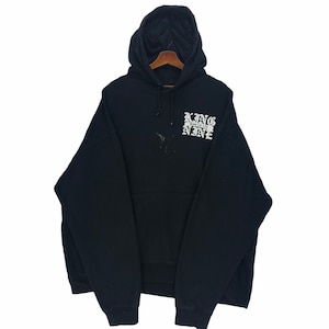 Può includere: Felpa con cappuccio nera con la scritta bianca "KING NINE", con cappuccio con coulisse e tasca frontale. La felpa è esposta su una gruccia di legno su sfondo bianco. L'indumento ha uno stile casual.