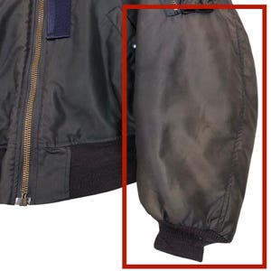 May include: Close-up of a dark green bomber jacket sleeve. The sleeve is made of a smooth, slightly shiny material and has a black ribbed cuff. A gold zipper is visible on the jacket's body.