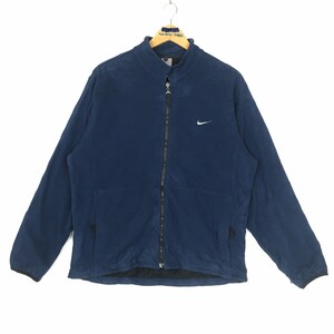 Puede incluir: Chaqueta azul marino con cremallera y un pequeño logotipo blanco de Nike en el pecho. La chaqueta tiene cuello alto y dos bolsillos delanteros. La chaqueta está colgada en una percha de madera.