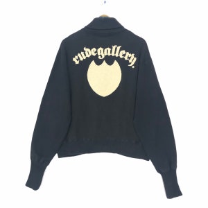 Op de afbeelding: Zwart sweatshirt met een hoge kraag en lange mouwen. De achterkant heeft de tekst "rudegallery" boven een crèmekleurig schildvormig ontwerp. Het sweatshirt hangt aan een houten kleerhanger.