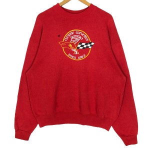 Puede incluir: Una sudadera roja con cuello redondo y mangas largas. La parte delantera presenta un gráfico circular con las palabras "Cyclone Corvettes Ames Iowa" que rodea un tornado, una bandera a cuadros y una bandera con una hoja.