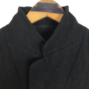 May include: Close-up of a black wool coat with a unique collar design. The coat is hanging on a wooden hanger. The coat's fabric appears to be a solid, dark color.