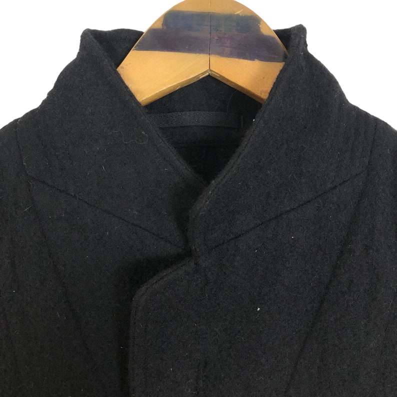 May include: Close-up of a black wool coat with a unique collar design. The coat is hanging on a wooden hanger. The coat's fabric appears to be a solid, dark color.