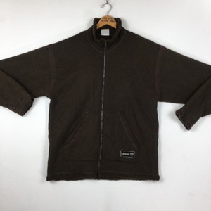 Puede incluir: Chaqueta marrón con cremallera, cuello alto y dos bolsillos delanteros. La chaqueta tiene una textura acanalada y una pequeña etiqueta en la parte inferior derecha. La chaqueta está colgada de una percha metálica.