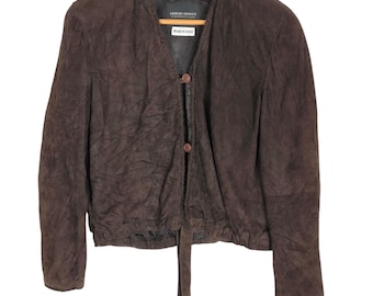 May include: A brown suede jacket with a V-neckline and three buttons down the front. The jacket has a tie at the waist and the label reads "GIORGIO ARMANI MADE IN ITALY". The jacket is on a wooden hanger.