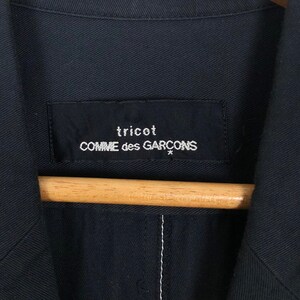 May include: Close-up of a navy blue jacket with a label that reads "tricot COMME des GARCONS" in white text. The jacket is hanging on a wooden hanger. The stitching is visible on the jacket.