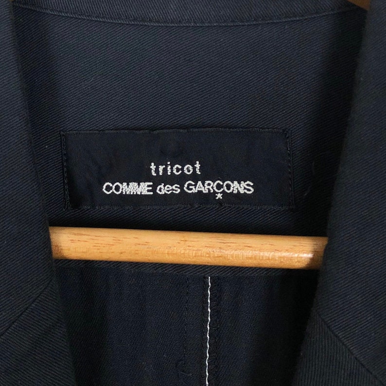 May include: Close-up of a navy blue jacket with a label that reads "tricot COMME des GARCONS" in white text. The jacket is hanging on a wooden hanger. The stitching is visible on the jacket.