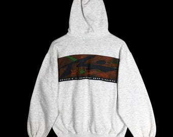 May include: A light gray hoodie with a hood and long sleeves. The front features a horizontal brown and green camouflage design with the text "RUSTY V. SURFBOARDS" in white.