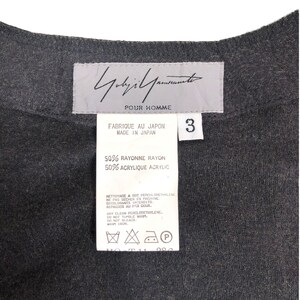May include: Close-up of a dark gray garment tag with the brand "Yohji Yamamoto POUR HOMME" printed in black. The tag also includes fabric composition details and care instructions. The number "3" is visible.