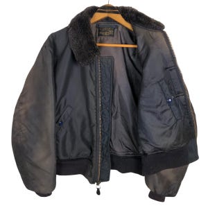 May include: A vintage black bomber jacket with a faux fur collar. The jacket features a zipper closure, multiple pockets, and ribbed cuffs and hem. The jacket is hanging on a wooden hanger.